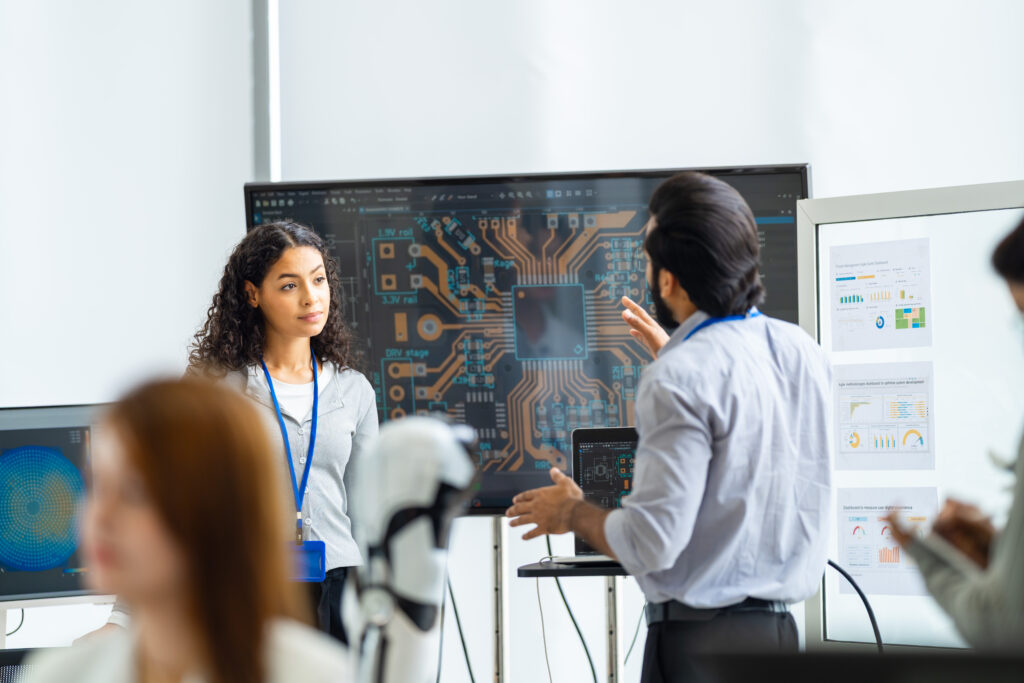 In a glass-walled innovation office, curly-haired female engineer with lanyard gestures at a large circuit board layout screen as bearded colleague explains, in an authentic workplace scene.